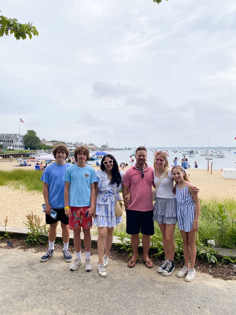 Fourth of July in Nantucket: A family of six poses in front of a beach. The beach behind them is busy with people and boats in the water. The sky is cloudy, and the family is dressed in casual summer clothing.