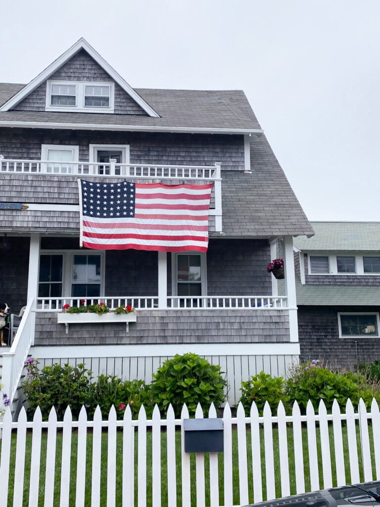 Fourth of July in Nantucket A two-story gray shingled house with a large American flag hanging on the front balcony, a white picket fence, and a small garden with flowers and shrubs.