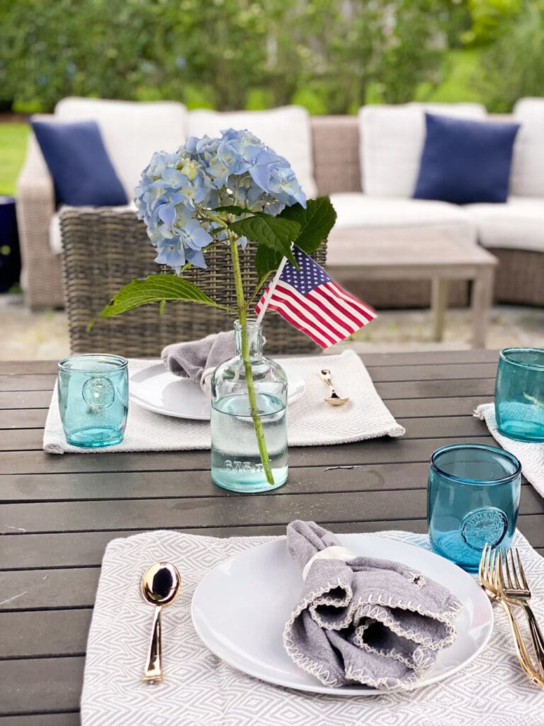 Fourth of July in Nantucket: Outdoor table setting with two place settings featuring blue hydrangeas in a vase with a small American flag, blue glasses, and grey napkins on a wooden table. Cushioned seating in the background.