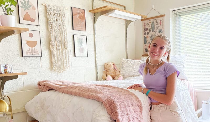 A young woman stands in a well-decorated bedroom with art on the walls, a teddy bear on the bed, and a window bringing in natural light. She smiles while leaning on the bed, which has a pink blanket.