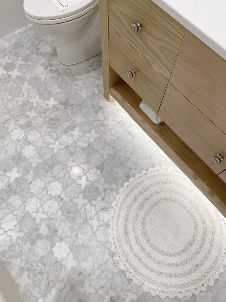 2025 Bathroom Trends Bathroom with white and gray patterned tile floor, wooden vanity with drawers, a round white rug, and a toilet in the background.