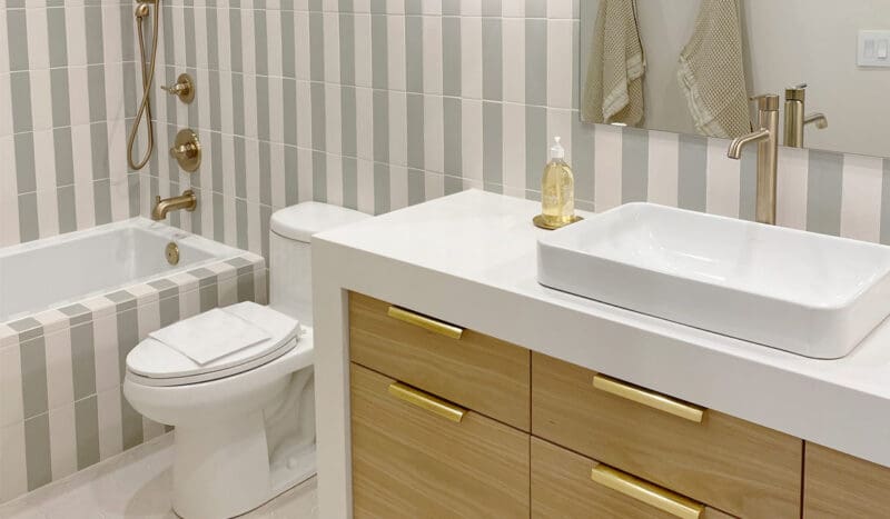 Bathroom with striped tile walls, a bathtub, toilet, and modern sink on a wooden vanity. There's a gold faucet and soap dispenser, and towels hang near the mirror.