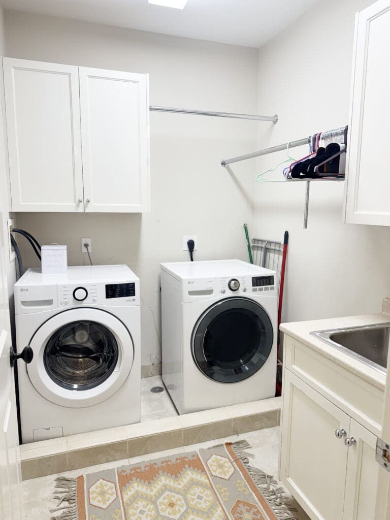 Laundry room with a white washer and dryer, cabinets, a sink, clothes rack, and cleaning tools. A patterned rug is on the floor.