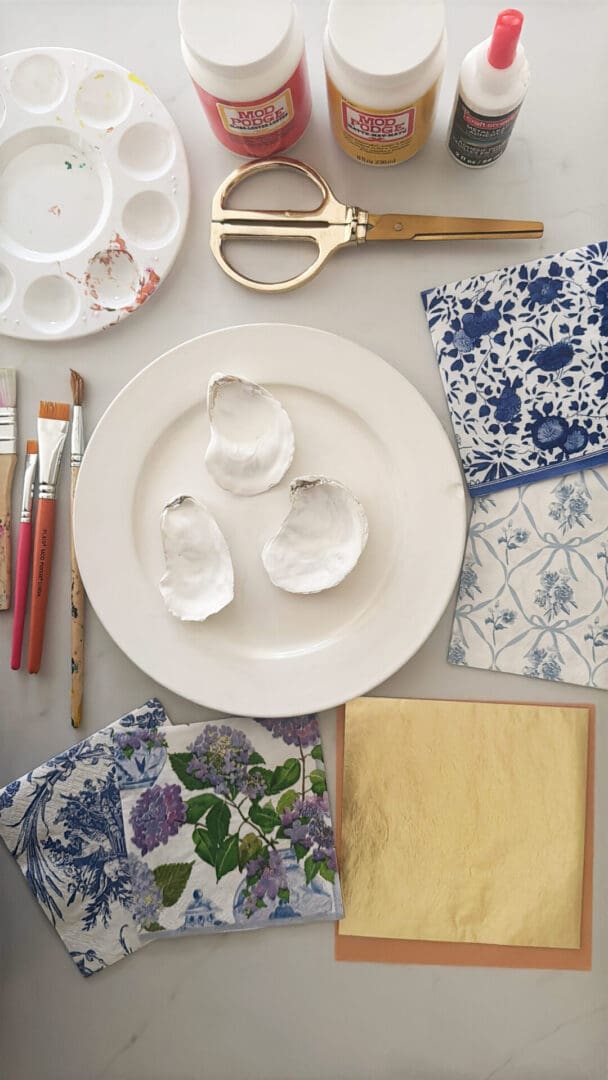 Crafting supplies arranged on a table: paint palette, brushes, gold leaf, scissors, Mod Podge, glue, decorative napkins, and oyster shells on a white plate.
Easy DIY oyster shell ring dish using paper napkins that is a beautiful gift to give for Easter or Mother's day or make for yourself. 