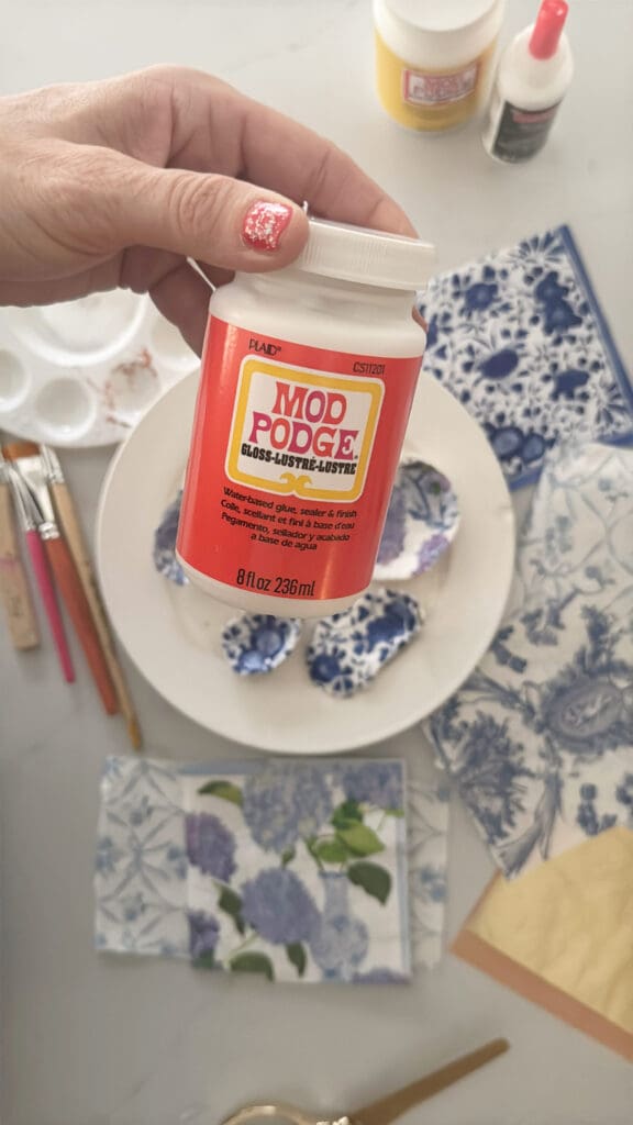 Hand holding a container of Mod Podge above a table with decorative papers, paintbrushes, and oyster shells.