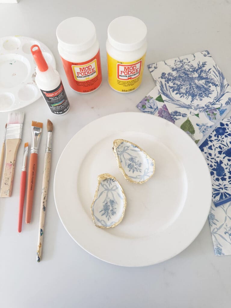 Art supplies and decorative items on a table, including Mod Podge jars, paintbrushes, glue, floral napkins, and gilded shells on a plate.
Easy DIY oyster shell ring dish using paper napkins that is a beautiful gift to give for Easter or Mother's day or make for yourself. 