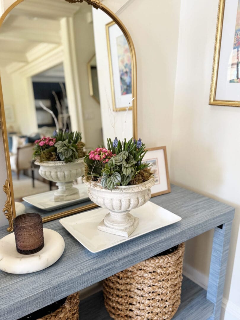 A decorative urn with pink and purple flowers sits on a tray atop a blue console table with woven baskets below, next to a round candle holder and a large mirror reflecting the room.