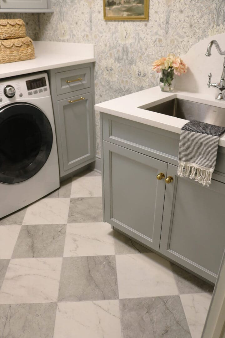 Laundry Room Renovation: Before and After 
Darling Darleen || Top CT Lifestyle Blogger A laundry room with gray cabinets, a front-loading washing machine, a stainless steel sink, checkered tile floor, and floral wallpaper. A towel hangs by the sink.
