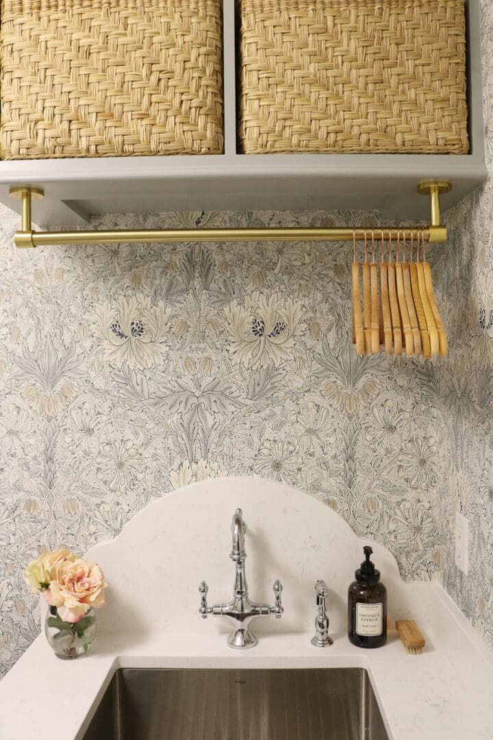 Laundry Room Renovation: Before and After 
Darling Darleen || Top CT Lifestyle Blogger
A laundry room sink with a marble backsplash, silver faucet, soap dispenser, scrub brush, flowers in a vase, woven baskets, and a gold hanging rod with wooden hangers above.
