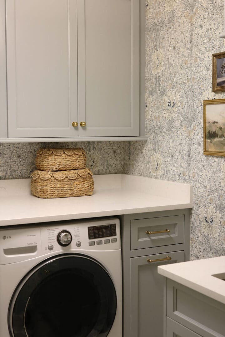 Laundry Room Renovation: Before and After 
Darling Darleen || Top CT Lifestyle Blogger
A laundry room with light gray cabinets, a front-loading washing machine, stacked woven baskets on the counter, and floral wallpaper with framed pictures on the wall.
