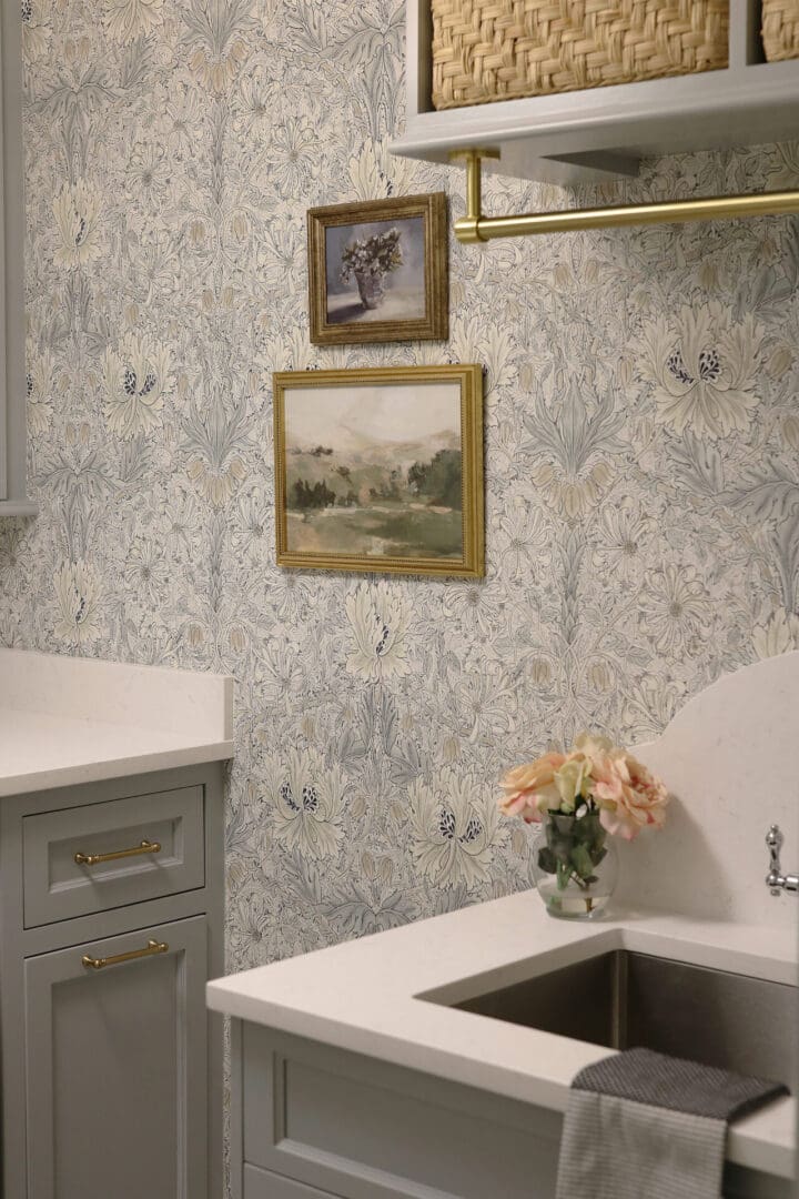 Laundry Room Renovation: Before and After 
Darling Darleen || Top CT Lifestyle Blogger
A laundry room with floral wallpaper, two framed paintings, a countertop with a sink, a small vase of flowers, cabinets, and a woven basket on an overhead shelf.