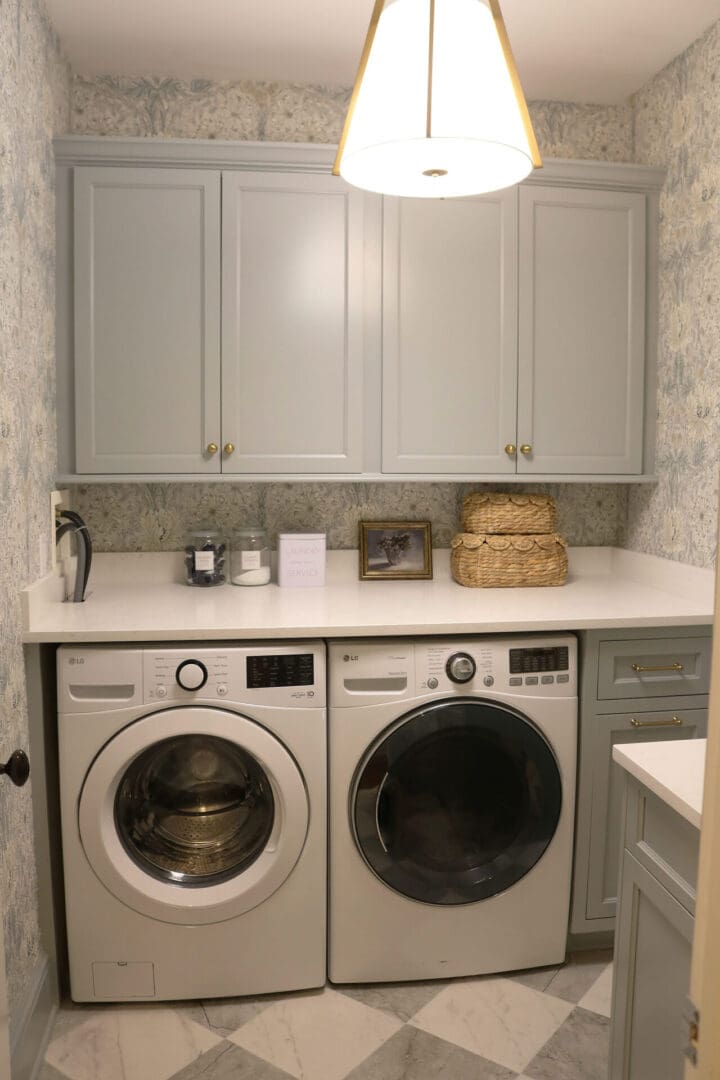 Laundry Room Renovation: Before and After 
Darling Darleen || Top CT Lifestyle Blogger A laundry room with front-loading washer and dryer, light gray cabinets, a countertop with baskets, a framed photo, a candle, and patterned wallpaper.