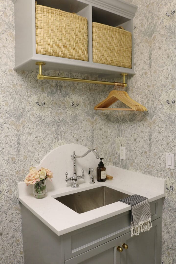 Laundry Room Renovation: Before and After 
Darling Darleen || Top CT Lifestyle Blogger Laundry room sink area with a silver faucet, a vase of flowers, soap dispenser, striped towel, wooden hangers on a brass rod, and woven baskets on a shelf above.