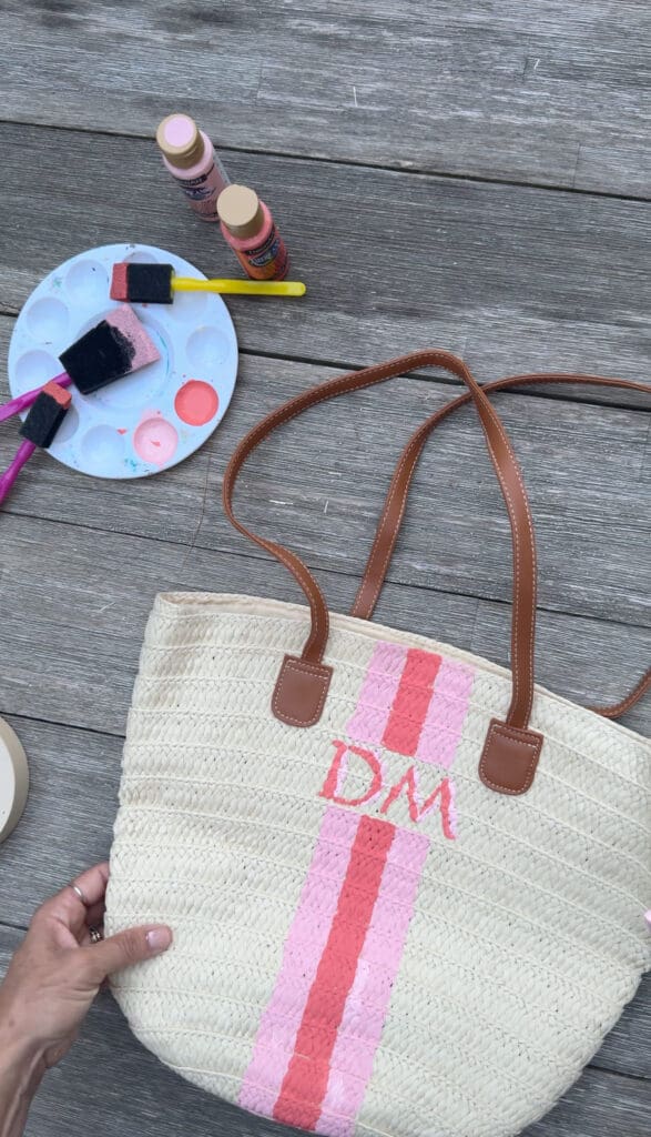 A hand holds a straw tote bag with brown handles, painted with pink stripes and the letters "DM." Nearby are paint supplies on a wooden surface.