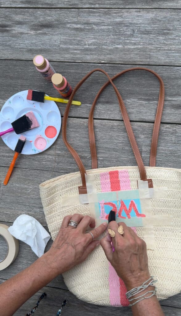 A person paints pink and blue letters on a woven straw bag with brown handles, using masking tape, paint, and brushes on a wooden surface.