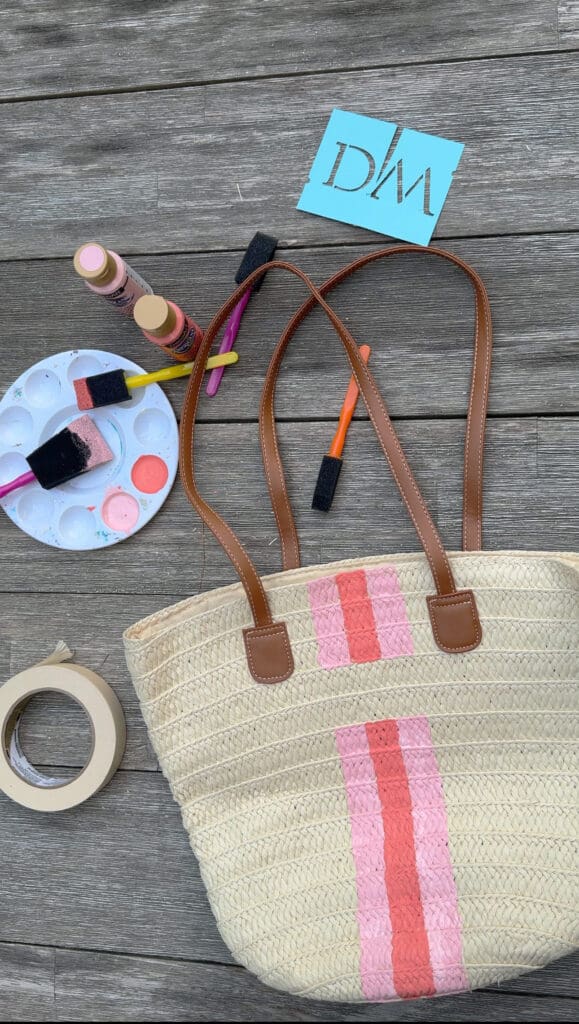 A woven tote bag with pink stripes is placed on a wooden surface, surrounded by paint supplies, a paint palette, tape, and a blue card with "DWA" printed on it.