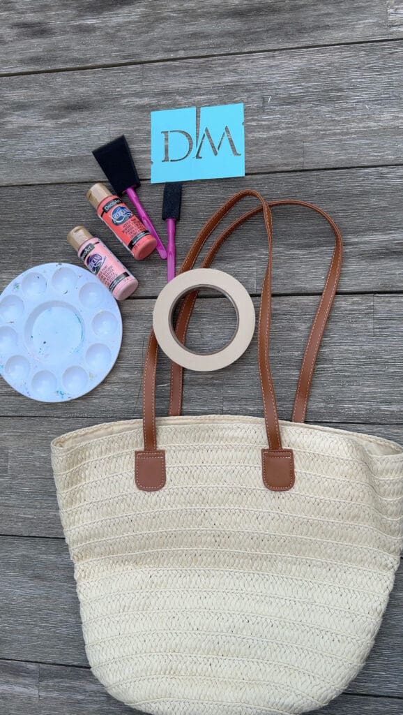 A straw tote bag, masking tape, paint palette, foam brushes, paint bottles, and a blue stencil with "DM" on a wooden surface.