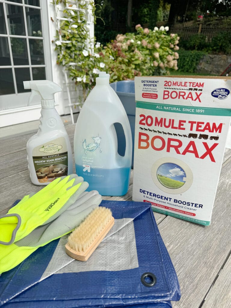 The simple tasks of how to clean outdoor cushions requires just a little elbow grease and a sunny afternoon and you can have your outdoor cushions looking new Cleaning supplies on a wooden table, including borax, gloves, a brush, spray cleaner, detergent, and a blue bucket with plants and a window in the background.