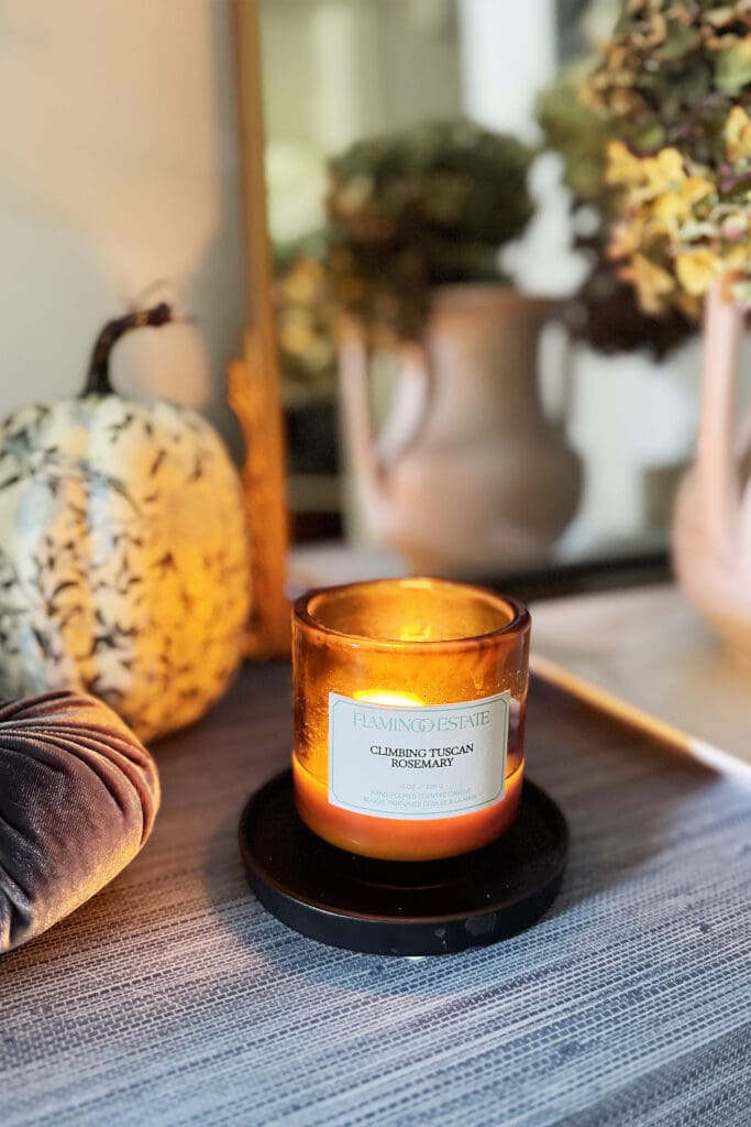 A lit candle in a glass jar sits on a black coaster next to a decorative pumpkin, with vases and flowers in the background.