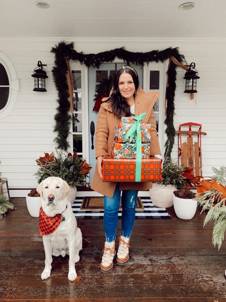 A woman in a brown coat holds a stack of wrapped gifts on a porch decorated for the holidays. A dog wearing a red scarf sits beside her.